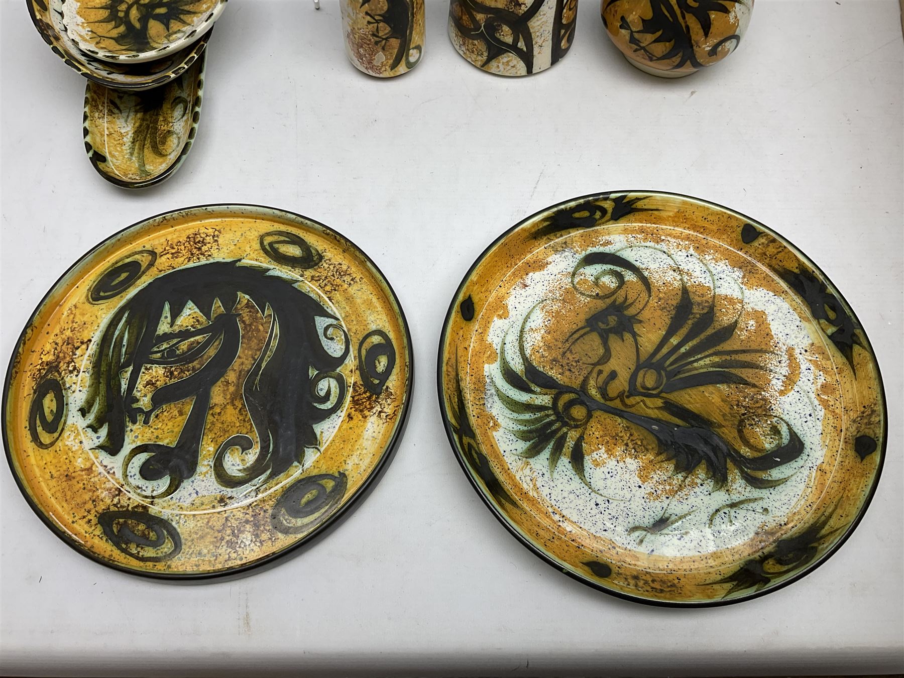 20th century Celtic Pottery, Newlyn, vases of baluster and cylindrical form, ovoid shaped bowl, plates, rectangular shallow dish etc, all decorated with a dark stylised phoenix, modelled with wings outstretched upon sponged ochre, blue and cream ground, together with a plate decorated with a stylised horse and tankard of tapering form (8)