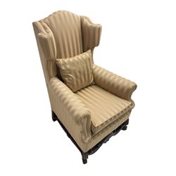 William and Mary style wingback armchair, upholstered in gold striped fabric, on walnut scroll carved supports joined by scroll and foliate carved middle rail, turned H-shaped stretchers, on castors