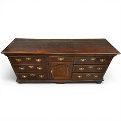 19th century Georgian oak long dresser base, rectangular plank top above a central frieze drawer over a fielded panel cupboard, flanked by three banks with quarter cantered pilasters, brass swan neck handles and escutcheons, raised on shaped bracket feet