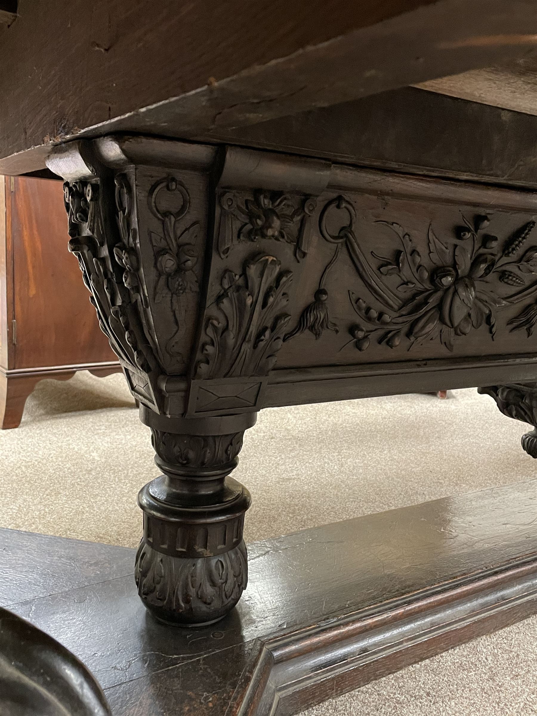 Large 19th century carved oak library table, the rectangular top with lunette carved edge over cartouche panelled and carved  scroll frieze, the central tapering rectangular support relief carved with masks and garlands, on four carved seated lion supports at each corner, on flat angular stretcher with recessed castors stamped ‘Bartholomew & Fletcher, London’