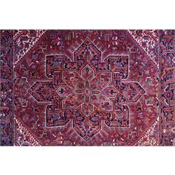 North West Persian Heriz hand-knotted carpet, brick-red field with a large central angular star medallion with pendant motifs, surrounded by stylized geometric palmettes, serrated leaves and tribal devices, ivory corner spandrels similarly decorated, field enclosed by a dark indigo main border of stylized rosettes and angular vine motifs between multiple guard stripes, with cotton fringe ends  