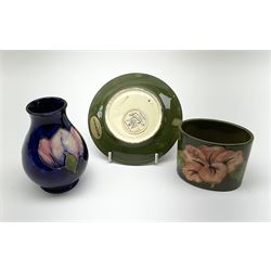 A small Moorcroft vase, decorated in the Magnolia pattern upon a dark blue ground, with impressed mark beneath, H9.5cm, together with two further pieces of Moorcroft, an oval pot and circular pin dish decorated in the hibiscus pattern upon a green ground, with impressed marks and paper labels detailed 'Potters to the late Queen Mary', pot H6cm, dish D12cm