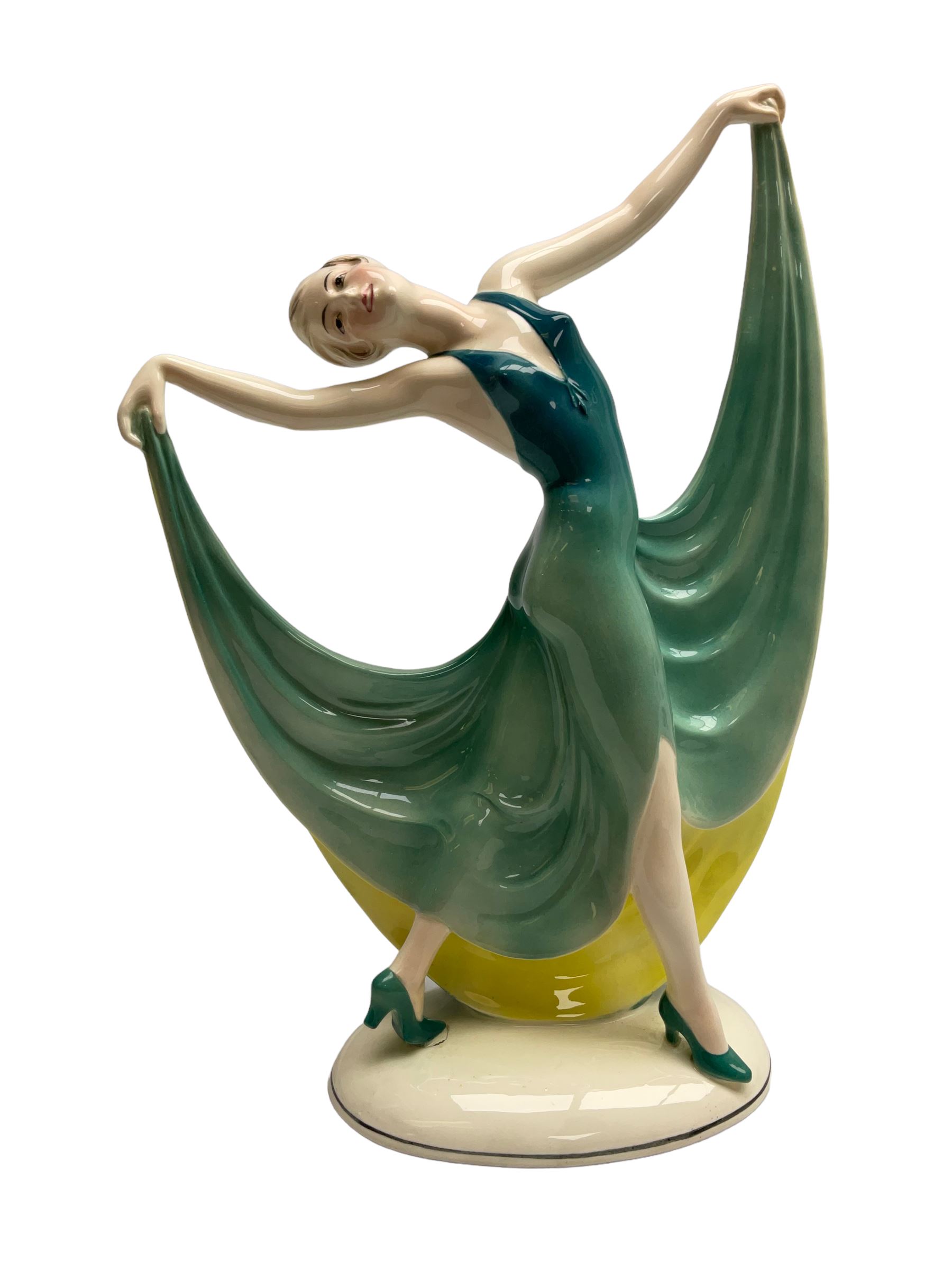 Early 20th century Hertwig Katzhütte figure of an Art Deco lady dancing, wearing a green, blue and yellow dress, stood upon on a domed plinth, with green cat H house printed mark beneath, H32cm