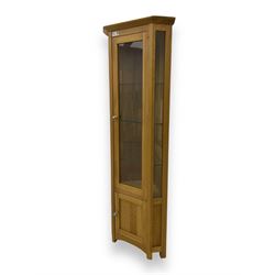 Contemporary light oak corner display cabinet, projecting cornice over single glazed door enclosing three glass shelves, the base fitted with panelled cupboard enclosing single oak shelf, on stile supports