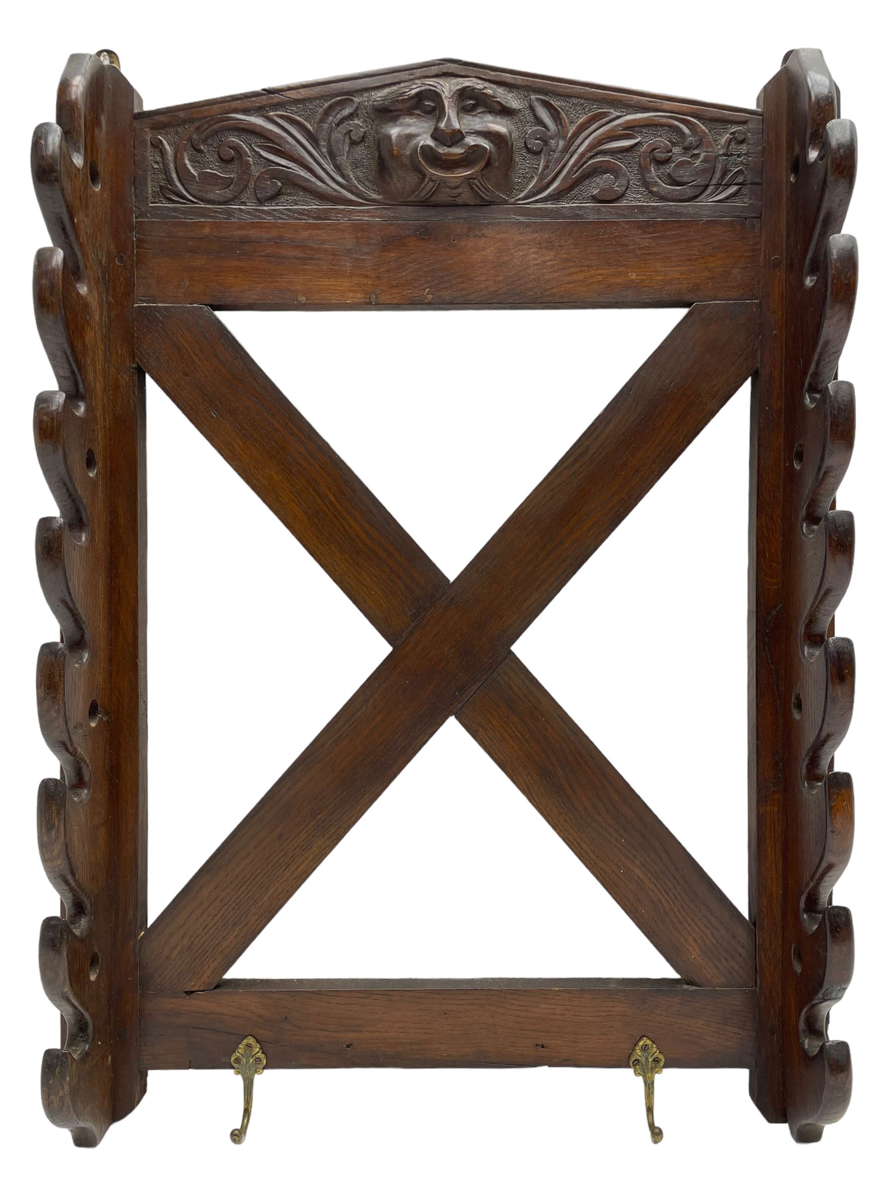 Victorian oak wall hanging whip or stick rack, sloped arch pediment carved with central grotesque mask with extended scrolling foliage, the upright supports cut with rests, x-framed back, two lower hooks