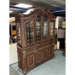 Kevin Charles American walnut display cabinet, illuminated interior, shell carved pediment above four bevel edge doors enclosing six glazed shelves above one central long drawer flanked by two short drawers, above three cupboards, carved bracket supports 