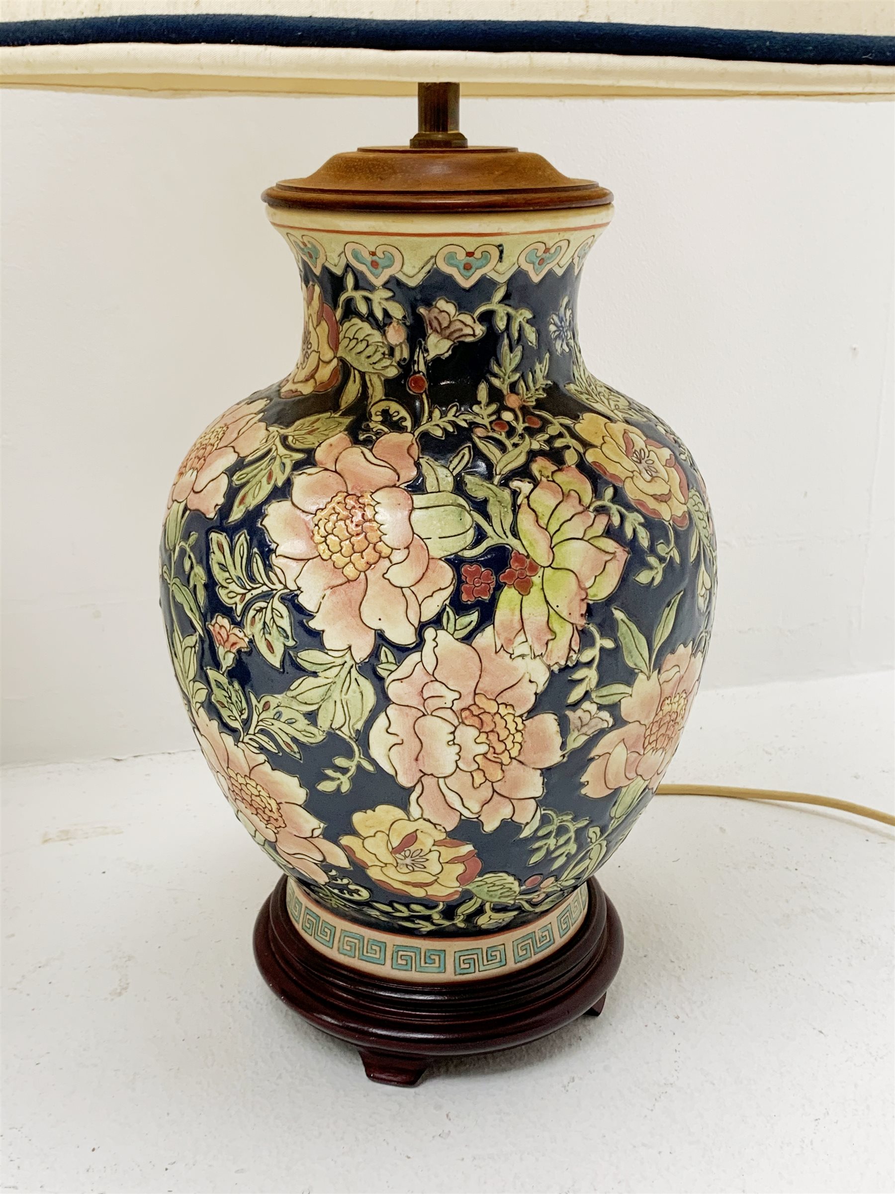 Two Oriental table lamps, each of bulbous form, the first example decorated with pink blossoming flowers upon a dark blue ground, above a fret pattern band, upon a wooden circular stepped base, the second example with white foliate decoration upon a dark blue ground, also with fret pattern band, and upon a wooden circular stepped base, each with fabric shade, including shade each approximately 74cm. 
