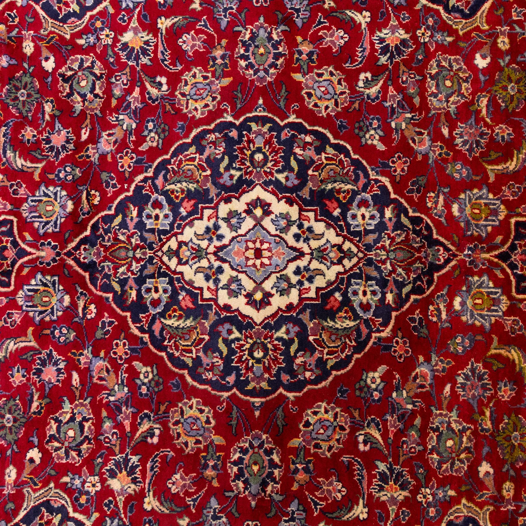 Central Persian Kashan hand-knotted rug, crimson red field with central ivory and indigo floral medallion surrounded by an all-over arrangement of finely drawn palmettes, flowering vines, and rosettes, with corresponding navy blue spandrels enclosed by a floral main border with multiple guard stripes, with cotton fringe ends    