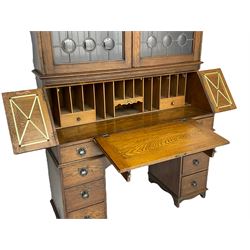 The Britisher Desk - early 20th century oak bureau cabinet, the raised cabinet enclosed by lead glazed doors, fall front with hinged side panels revealing fitted interior, twin pedestal base, fitted with nine drawers