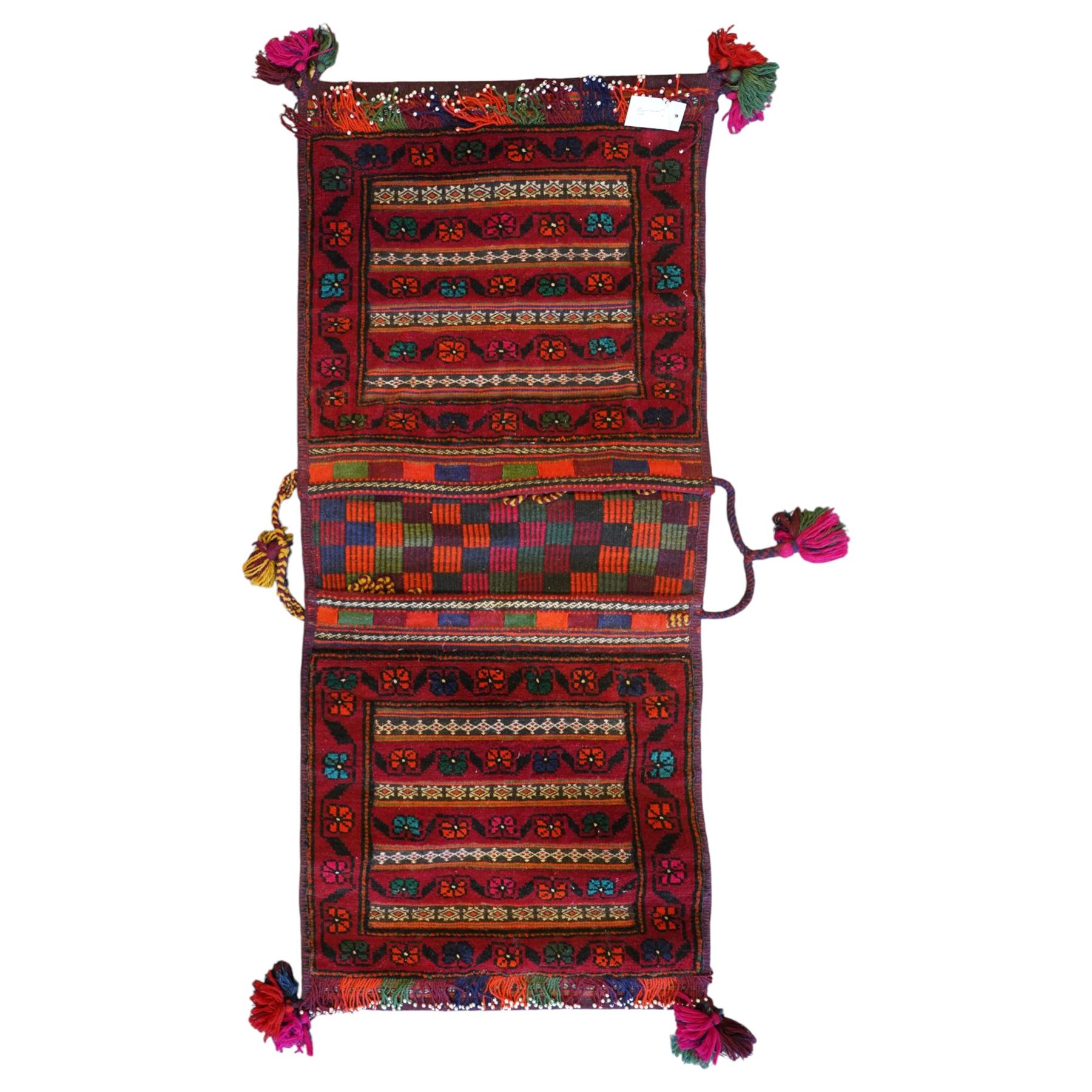 North East Persian Turkoman saddle bag, traditional double pouch form, each panel with madder red ground, decorated with horizontal bands of stylized geometric floral motifs and angular vine work, framed simple borders, central panel woven in flatweave with polychromed chequered design flanked by braided carrying cords with tasselled and knotted decorative elements 