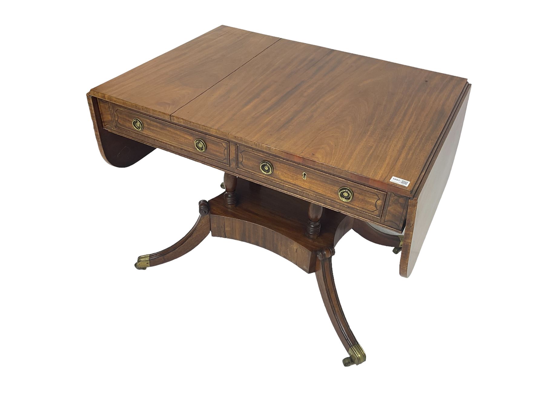 Regency period mahogany drop-leaf sofa table, rectangular top fitted with two cockbeaded drawers with ebony stringing, raised on turned columns united by quadrupod base, the sabre supports terminating in brass cups and castors