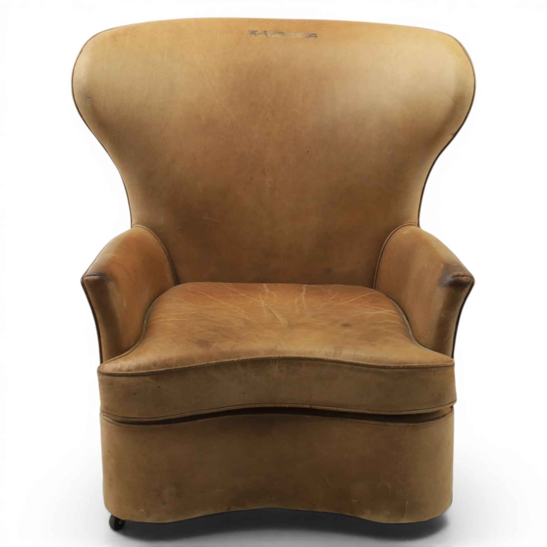 20th century tan leather armchair, the curved wing back above low square arms, shaped loose seat cushion with piped edge, close-nailed side detail, raised on concealed castors
