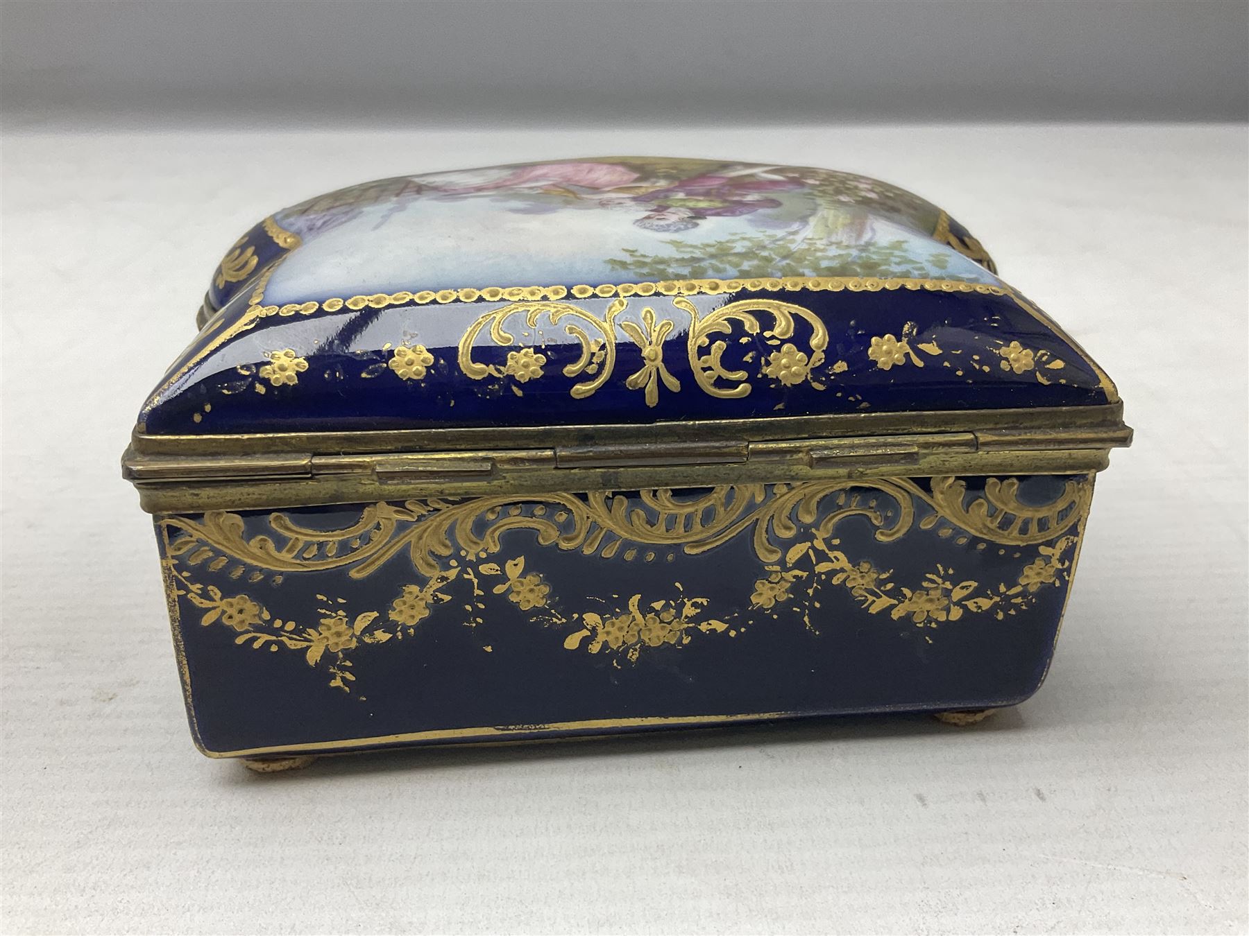 Sevres style late 19th/early 20th century trinket box decorated in the rococo style, the shaped cover painted with an 18th century courting couple in a garden scene opening to reveal the interior painted with floral sprays, the conforming body with a painted panel of floral decoration, all reserved upon cobalt blue ground with foliate gilt borders, with interlaced L's mark beneath, L12cm H6cm