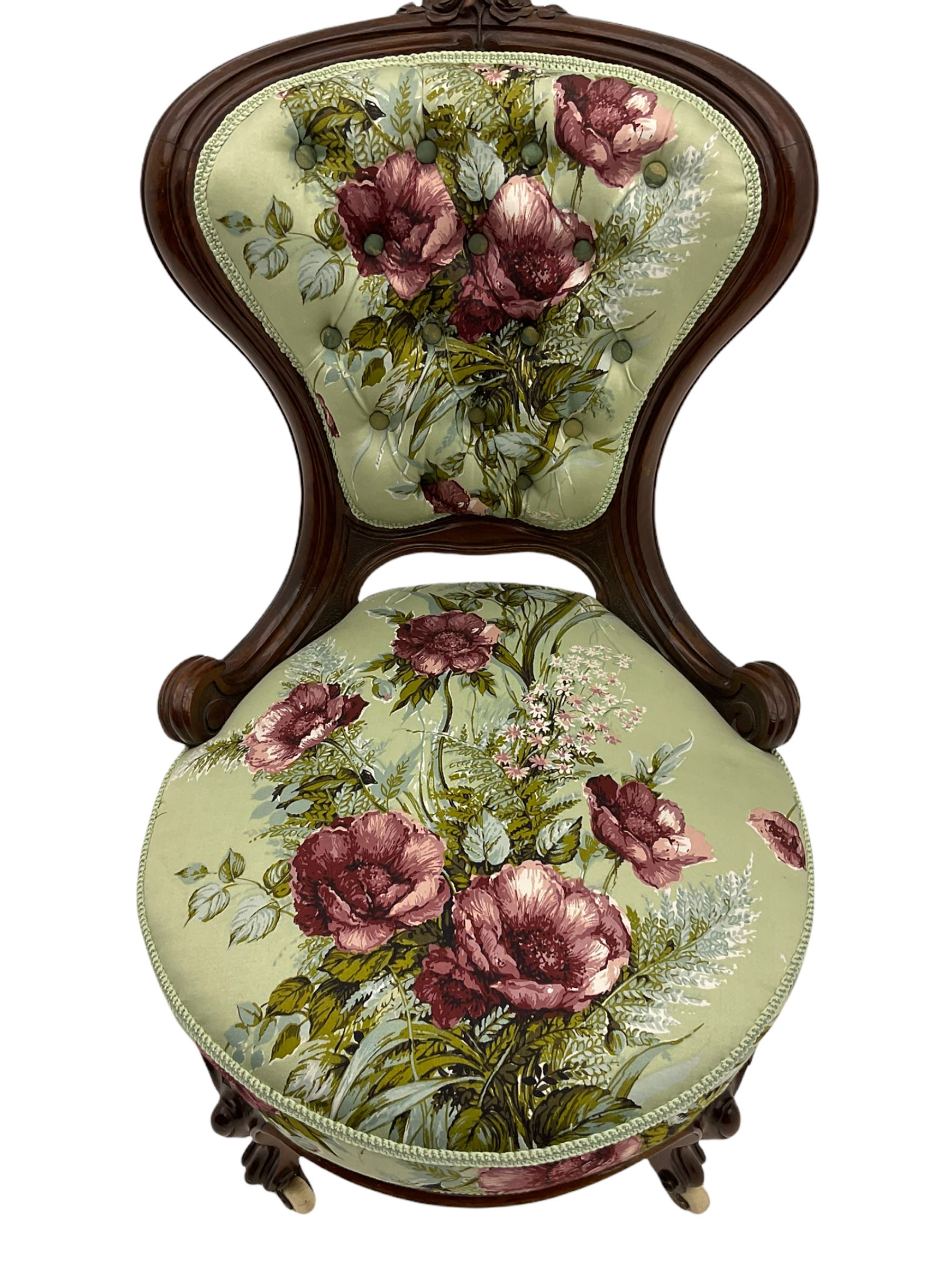 Victorian walnut framed nursing chair, spade shaped back carved with flower heads, upholstered in buttoned rose patterned fabric, on scroll carved cabriole supports terminating at brass and ceramic castors