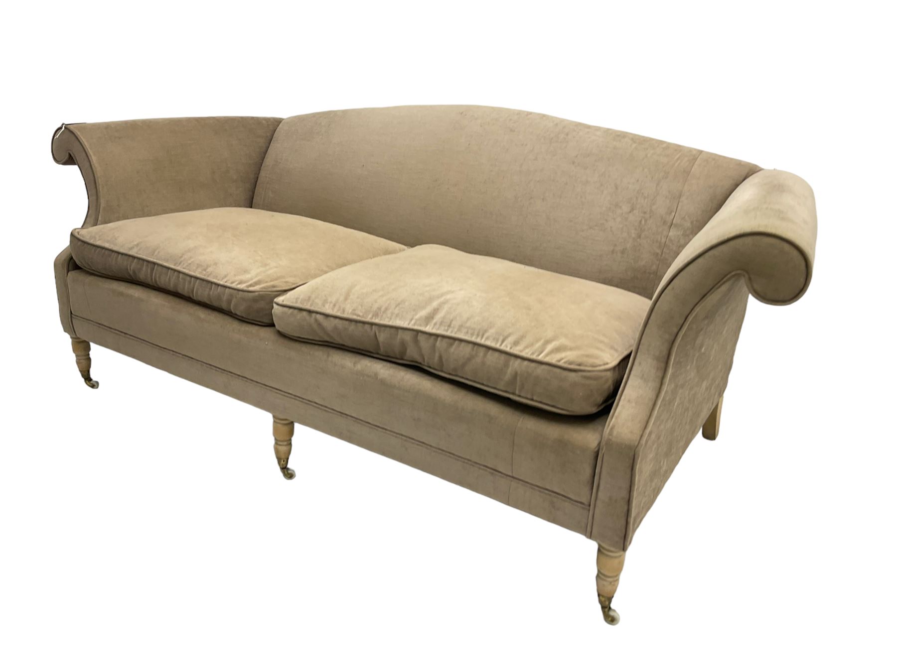 Traditional three seat sofa, curved back over scrolled arms, upholstered in crushed beige fabric with matching loose cushions, on turned front supports with brass and ceramic castors, together with matching footstool