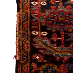 North West Persian Heriz hand-knotted carpet, madder red ground, with a large central geometric medallion with pendants, surrounded by angular foliate and floral motifs incorporating stylised rosettes, palmettes and serrated vine work, enclosed by ivory spandrels and a dark navy border with repeating geometric flowerheads and angular meandering vines, flanked by narrow patterned guard stripes, with cotton fringes