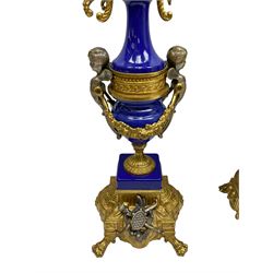 A 20th century continental gilt metal Lyre mantle clock with a pair of matching six light candelabra, spring driven movement housed in a gilt drum case on a brass and cobalt blue base with paw feet, two bronzed metal figures of mythological cherubs with animal legs and cloven hooves supporting a festooned garland swag, eight-day twin barrel striking movement with a floating lever balance escapement, striking the hours and half-hours on two bells, white enamel dial with roman numerals, minute track and pierced steel hands, dial inscribed “Imperial”. 