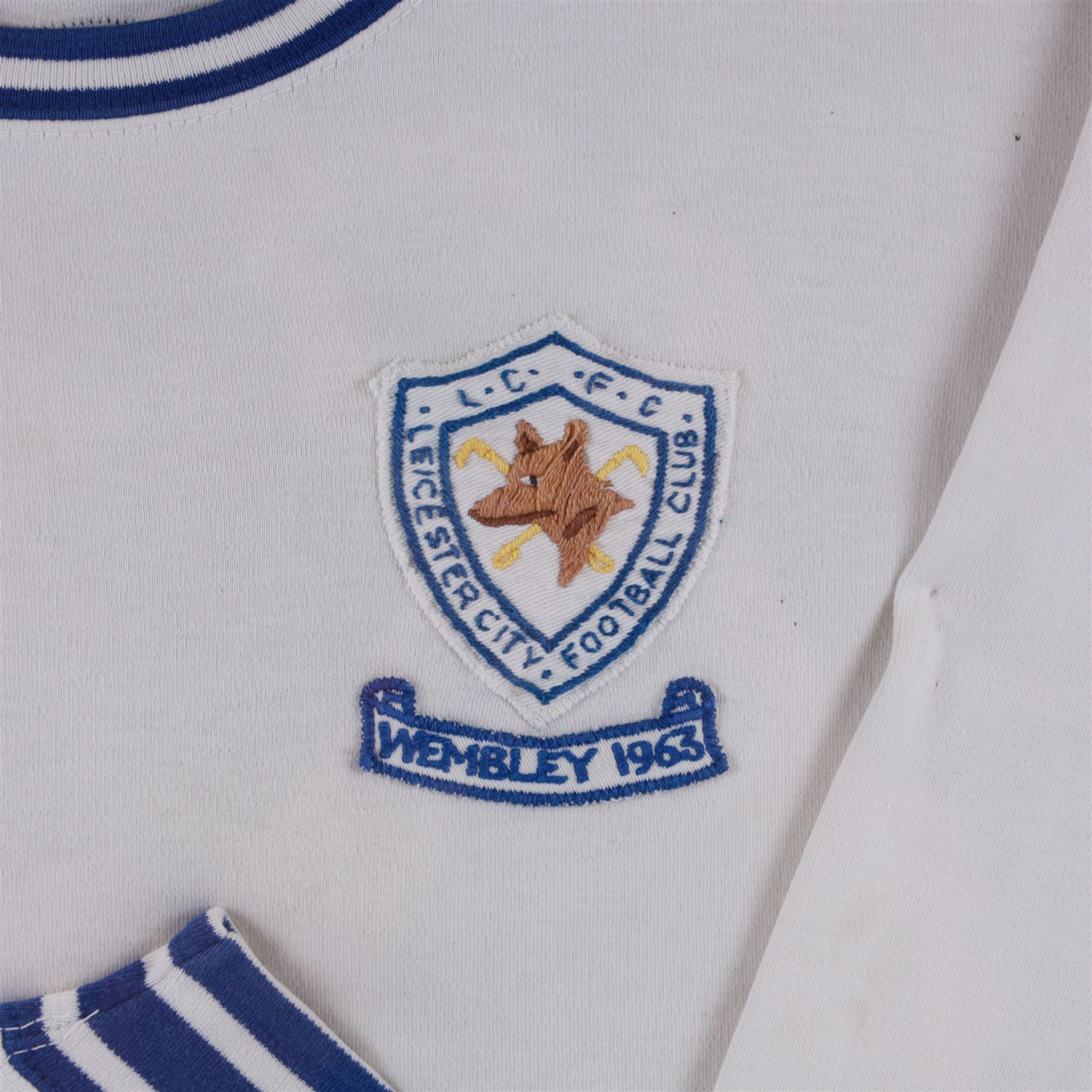 F.A. Cup Final Wembley 1963 Leicester City V Manchester United match worn long sleeved shirt, worn by Colin Appleton Leicester City captain, all white with striped blue collar and cuffs, embroidered cloth Leicester City badge with Wembley 1963 below, with Bukta Dyfast Zeebux label to inner collar, contained within a glazed wooden display frame with engraved plaque reading Colin Appleton (Capt) F.A. Cup Final Wembley 1963 Leicester City V Man Utd, frame H80cm

Notes: The 1963 F.A. Cup final was played on 25 May 1963 at Wembley 'Empire' Stadium. The final was won by Manchester United 3-1. 

Colin Harry Appleton (7 March 1936 – 31 May 2021) was an English footballer and manager. He captained Leicester during their 1962-63 season and the FA Cup final of this year. He then went on to captain them to the 1964 League Cup win. 

He later played for Charlton Athletic and Barrow, before playing for and managing Scarborough. He went on to manage Hull City, Swansea City, Exeter City and Bridlington Town.