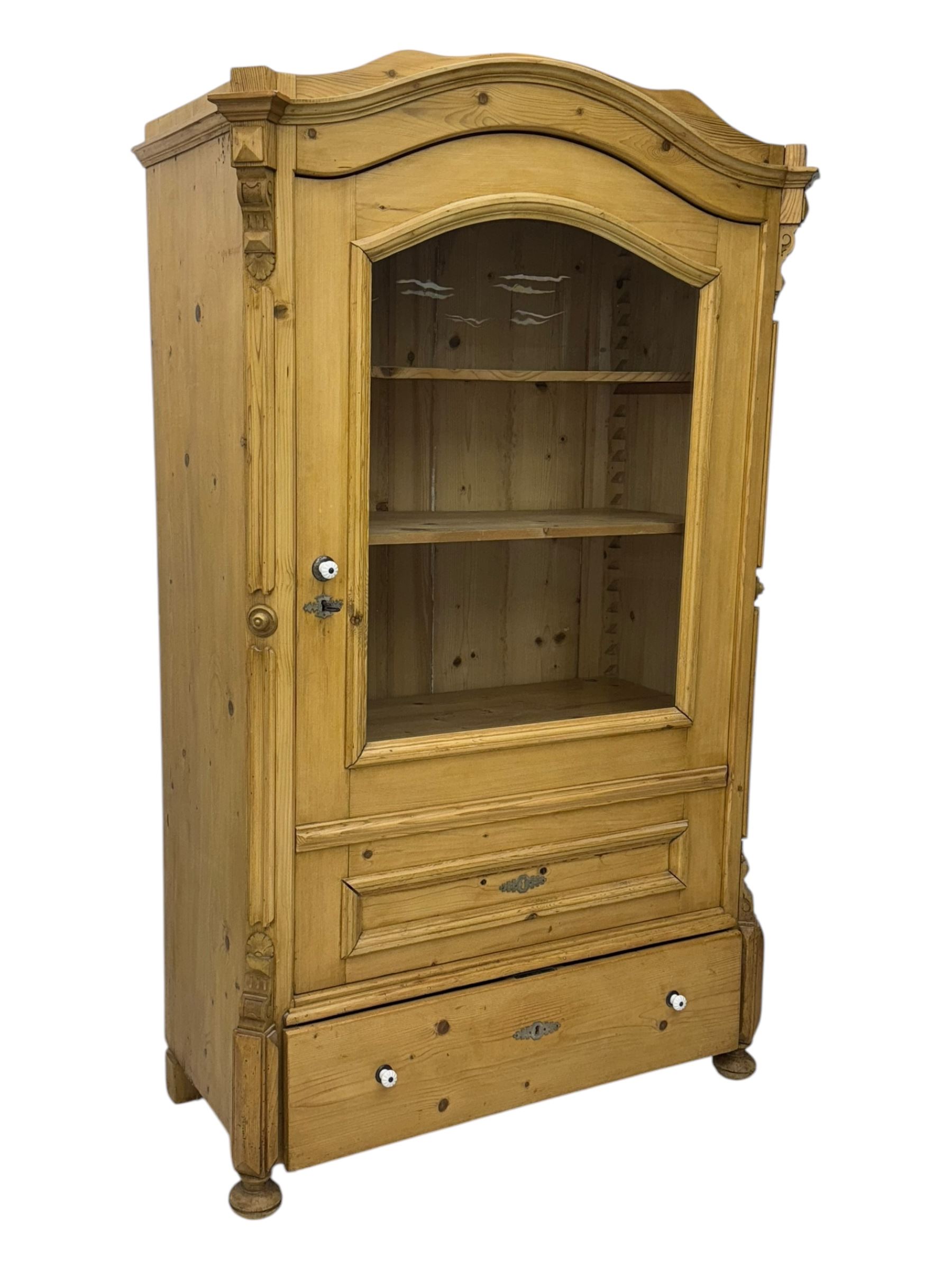 Late 19th century Continental pine vitrine, shaped and moulded arched cornice over a single glazed door enclosing adjustable shelves, flanked by carved pilasters with foliate and geometric motifs, fitted with single moulded drawer, raised on compressed bun feet