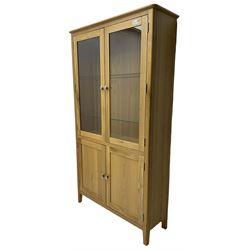Contemporary light oak display cabinet on cupboard, fitted with two glazed doors enclosing two adjustable glass shelves with light fittings to the top, over panelled cupboard concealing single shelf, on square supports