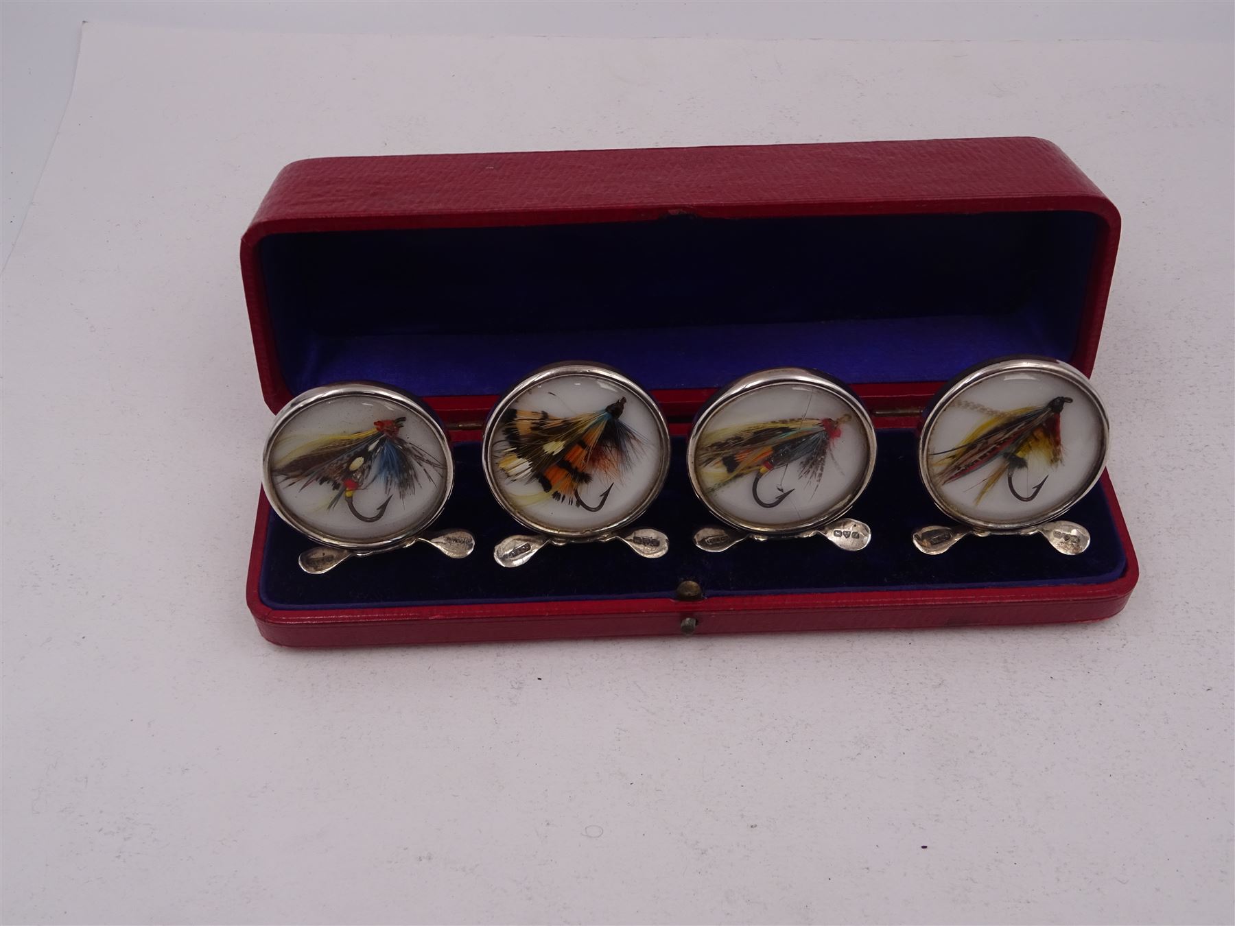 Set of four early 20th century silver mounted place card holders, each of circular form and set with a coloured fly fishing hook behind a glazed panel, upon three pad feet, hallmarked Grey & Co, Chester 1912, H4.5cm, contained within red fitted case, with purple silk and velvet interior