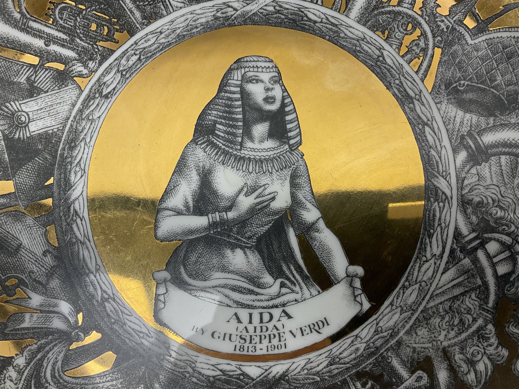 Three Fornasetti plates from the Melodramma series, each depicting a operatic portraits within a border of operatic emblems on gilt ground, with printed mark beneath, D25cm 