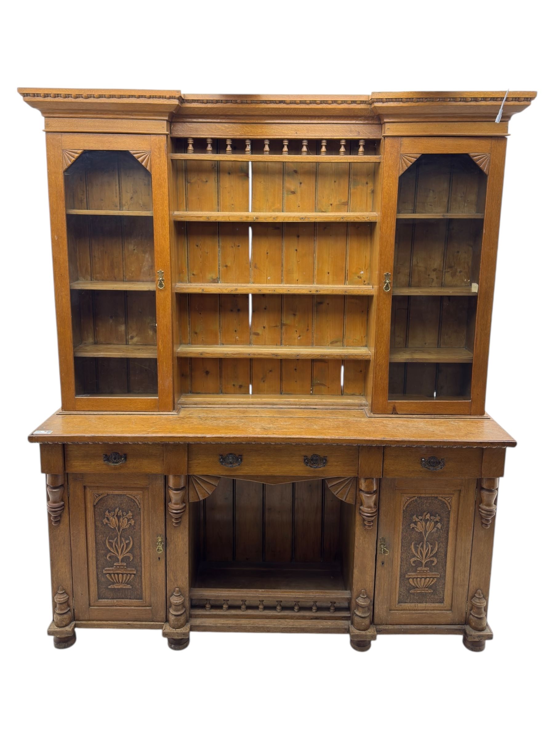 Late 19th century pine dresser, the projecting cornice with dentil and spindle moulded frieze above a three-tier plate shelves, flanked by glazed doors, the base fitted with three frieze drawers over arched apron and carved floral panelled cupboard doors, raised on turned feet