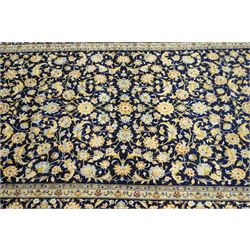 Central Persian Kashan hand-knotted carpet, deep indigo field with a densely all-over arrangement of scrolling floral vines, palmettes, and flowering sprays, in soft ivory, pale blue, and warm gold tones, principal ivory border with a continuous meandering vine of stylized palmettes, and floral motifs, flanked by multiple narrow guard borders, with cotton fringe ends 