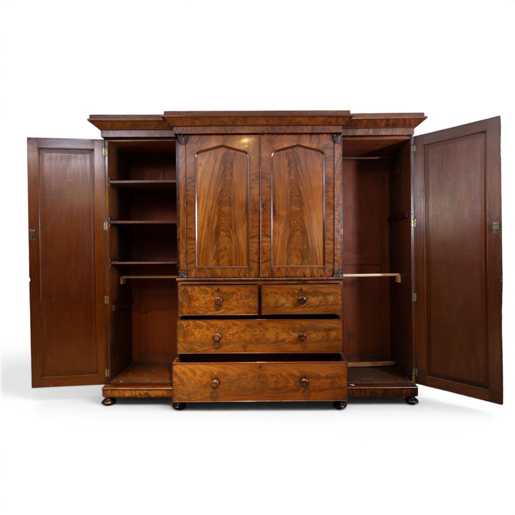 Early 19th century figured mahogany combination wardrobe, the breakfront architectural cornice over central press cupboard enclosing linen slides, flanked by full-height hanging cupboards with arched inset panels, lower arrangement of two short and two long drawers with turned knob handles, applied acanthus carved detail, raised on bun feet