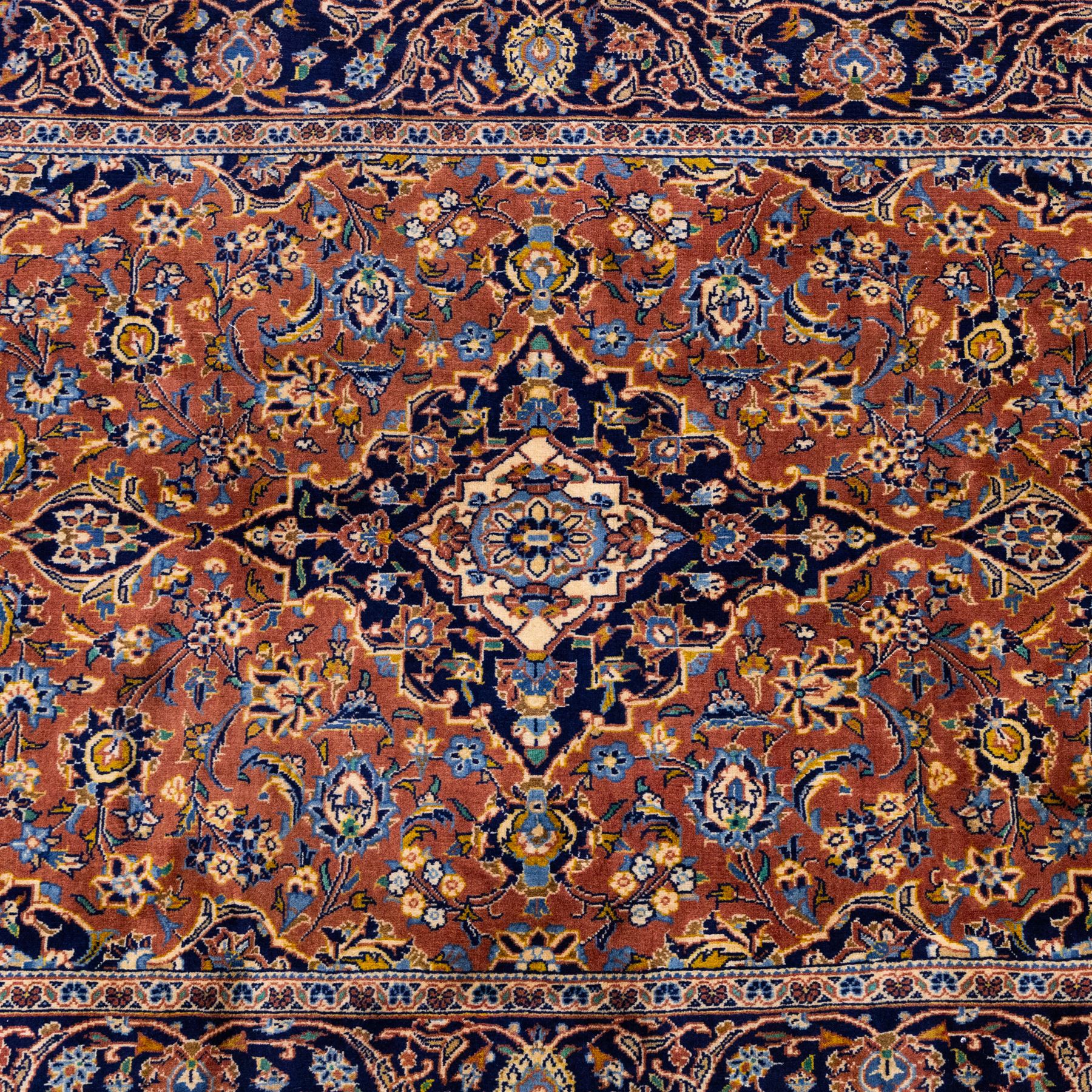 Central Persian Kashan hand-knotted rug,  terracotta field with central shaped ivory and dark blue medallion with palmettes, floral sprays and scrolling vine ornament, with conforming corner spandrels enclosed by a broad navy border of palmettes and flowering vine between multiple guard stripes, with cotton fringe ends 