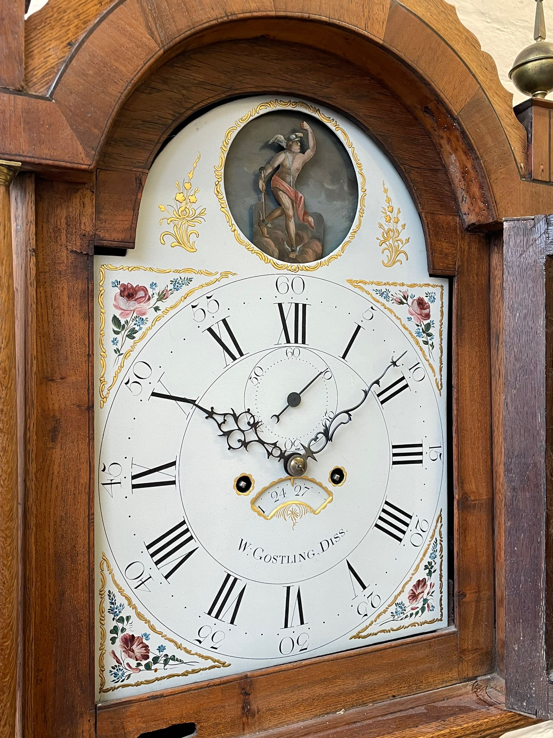 William Gostling of Diss  (Norfolk)-  Late 18th century 8-day oak longcase clock c1780-90, break arch top with scalloped cresting and three brass finials, glazed break arch hood door flanked by turned pilasters with brass capitals, trunk with a long trunk door and rounded top on a square plinth with an applied and shaped skirting, early painted Wilson of Birmingham dial with Roman numerals and five minute Arabic's, matching steel hands and second pointer, arched square ended calendar aperture with date disc behind, floral spandrels within gesso borders and a painted rocking automaton representing Mercury to the break arch, dial pinned via a false plate to a weight driven rack striking movement with a recoil anchor escapement, striking the hours on a cast bell. With pendulum weights and key. 
William Gostling (II) is recorded as working in Diss from 1774 to 1797, succeeded by widow Anne and son George . The dial of this clock is illustrated and discussed in detail in M.F. Tennants book 