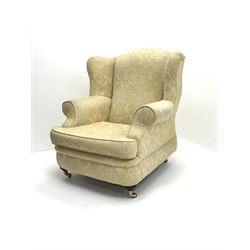 Queen Anne style wing back armchair, upholstered in pale golden floral patterned fabric, raised on turned supports and brass casters