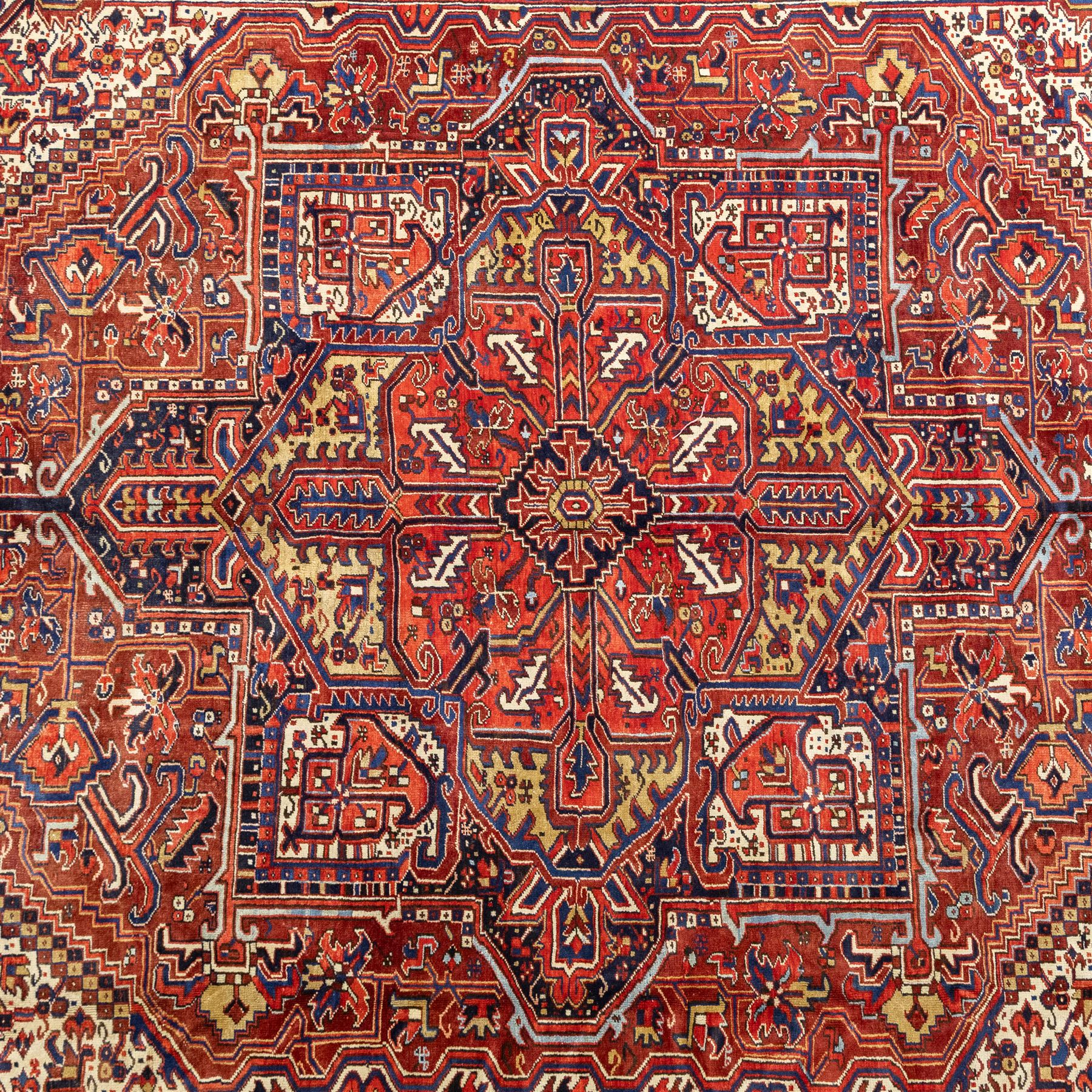 North West Persian Heriz hand-knotted carpet, madder red field, with a large angular stepped central medallion with serrated outlines, flanked by bold stylized palmettes, hooked leaves and geometric devices, spandrels similarly decorated within a wide geometric main border featuring angular floral motifs, enclosed by multiple guard borders in madder red, indigo blue, ivory, olive green and soft ochre, with cotton fringe ends 
