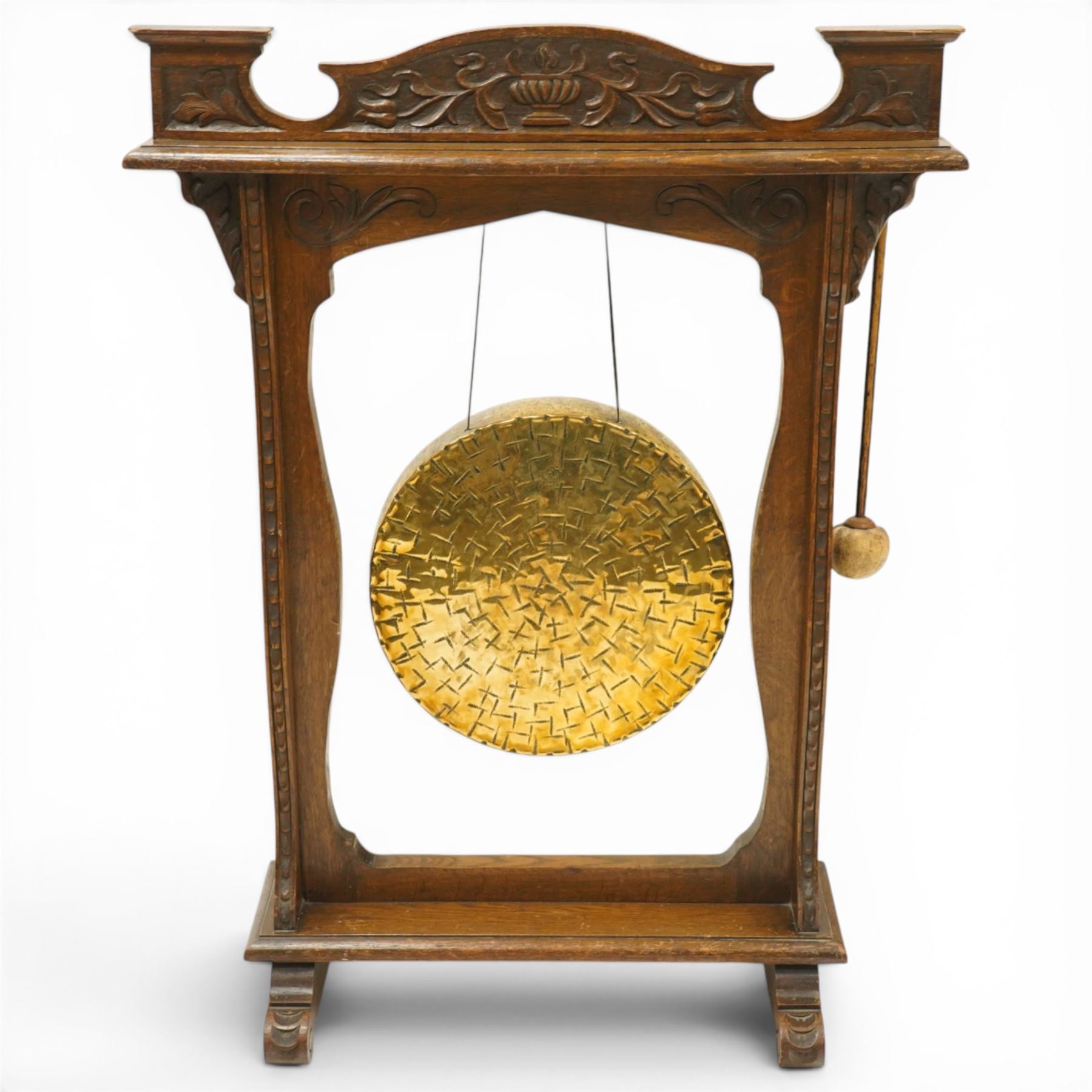 Late Victorian Arts & Crafts carved oak framed floor standing dinner gong, the cresting rail carved with central urn and trailing foliage, over shaped end supports decorated with graduating tulip and floral motifs, on rectangular base with scroll sledge feet, the brass gong stamped with William Suckling of Birmingham 'Burmese' makers mark, with striker
