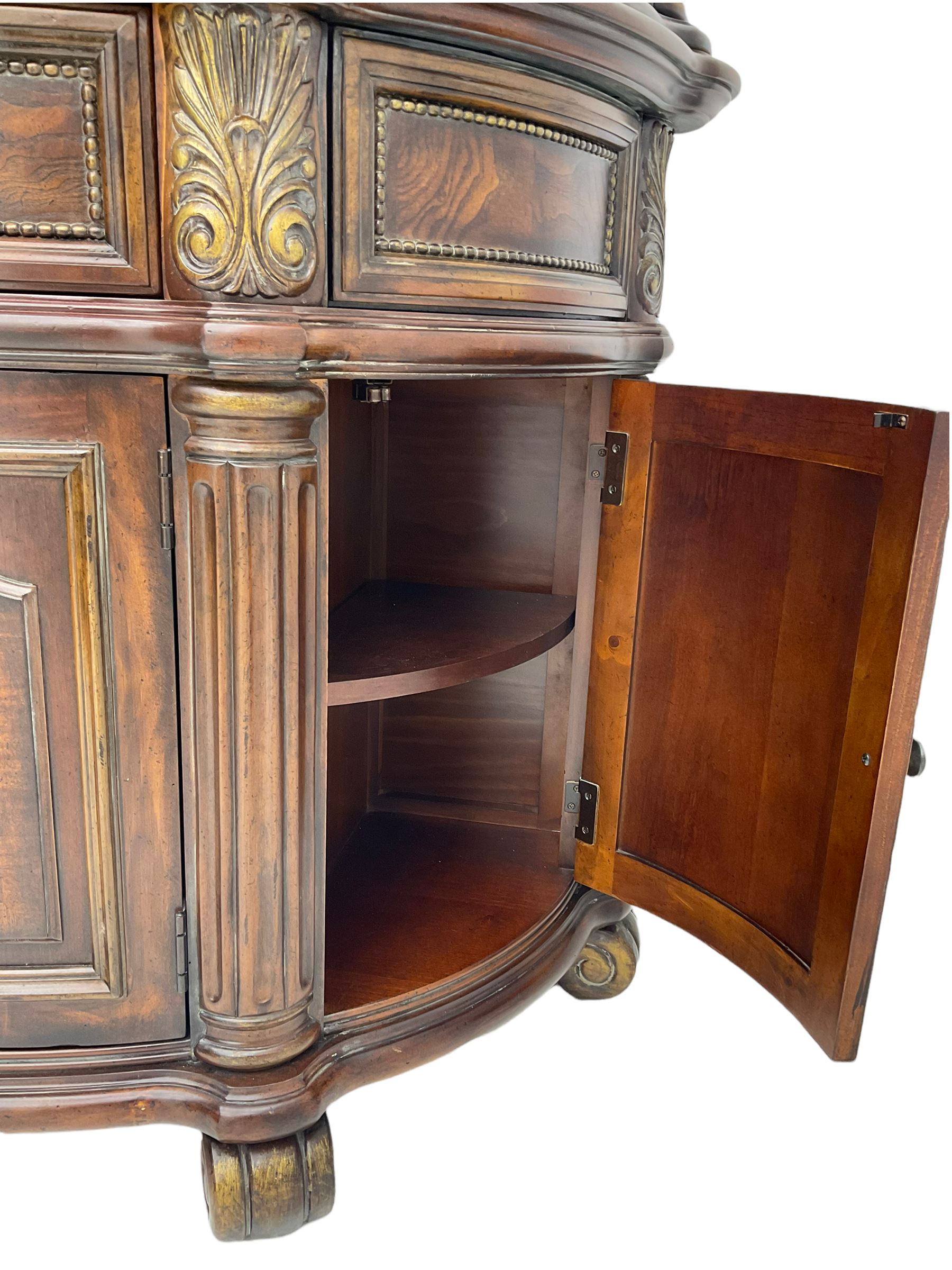  Kevin Charles American walnut demi lune display cabinet with illuminated interior, projecting cornice, two bevel edge glazed doors enclosing two adjustable shelves above single drawer and four cupboard doors with wine rack, carved scrolling supports