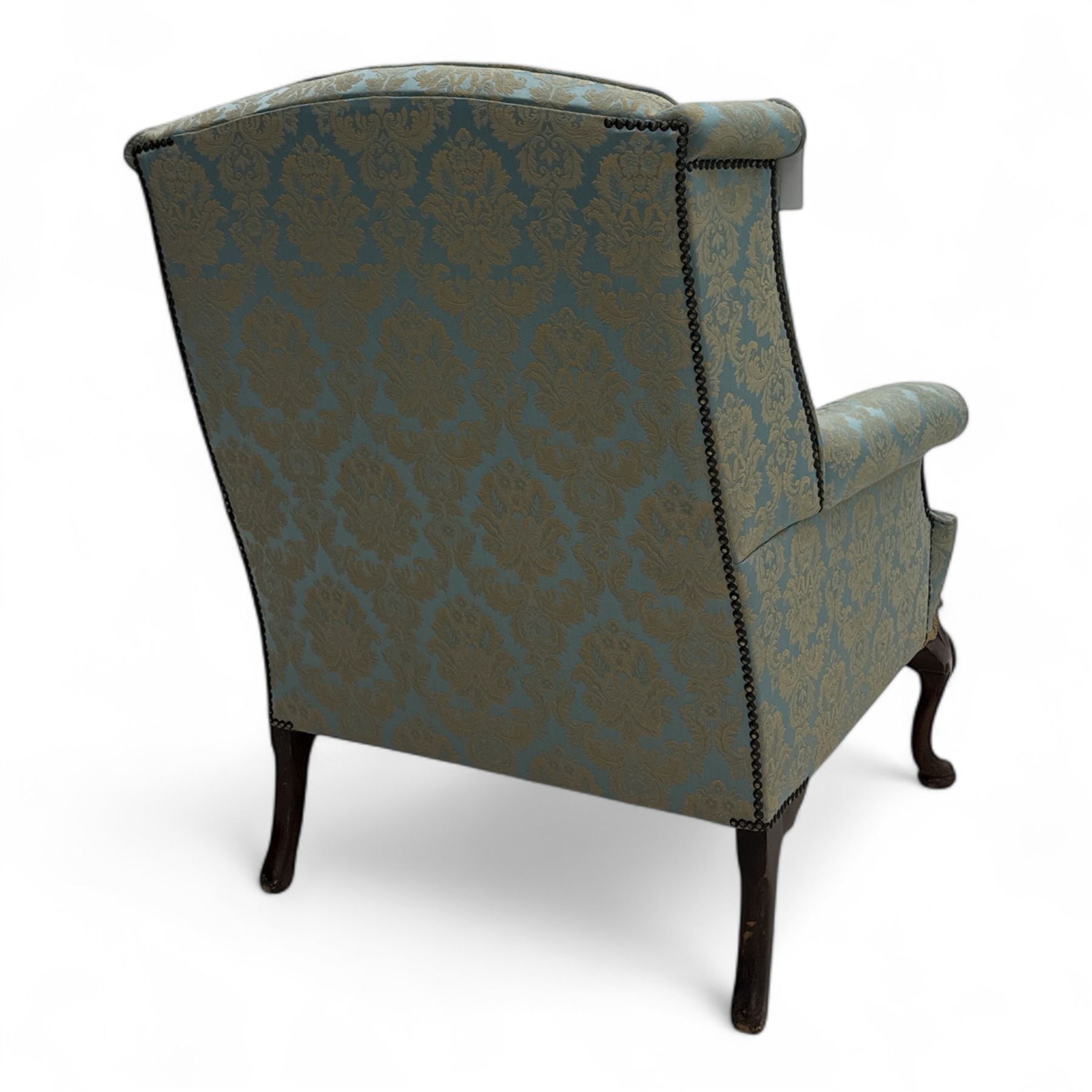 Georgian design carved walnut framed wingback armchair, upholstered in pale blue and gilt floral damask, with shaped loose seat cushion and rolled arms, raised on scallop and cartouche carved front cabriole supports terminating to pad feet