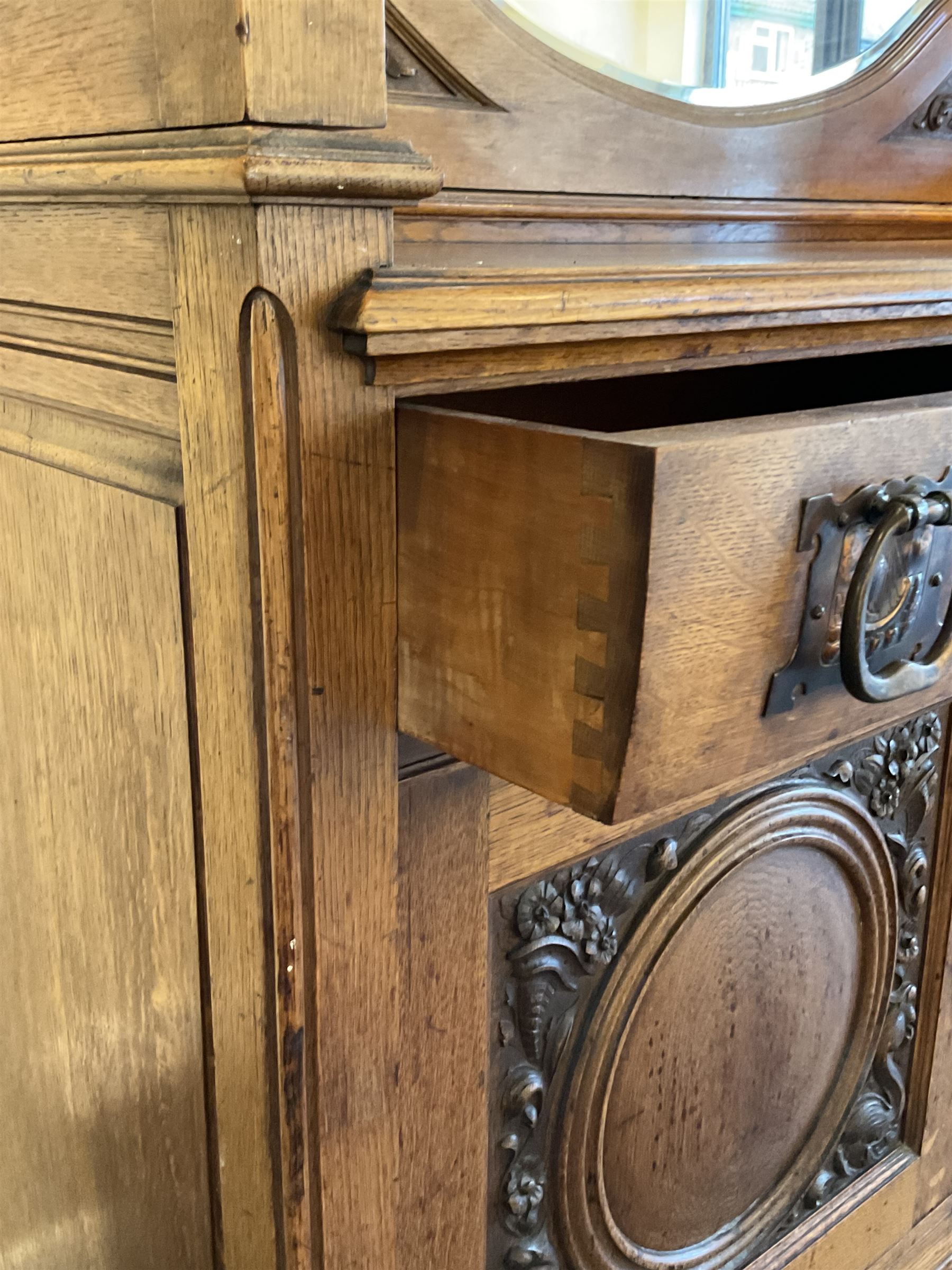 Late 19th century Arts & Crafts period oak mirror back sideboard, shaped bevelled mirror back carved with foliate scrolls and flower heads, down swept supports with elongated club-shaped uprights, moulded rectangular top over two drawers and three cupboards, the drawer fronts with copper handles decorated with sailing ships, panelled doors carved with mythical beasts, flowerheads and foliage, on moulded block feet