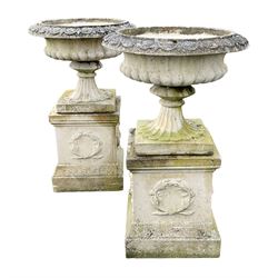 Pair composite stone urns on square plinths, the urns with egg and dart moulded rim over gadrooned body, fluted splayed support on square base, the square plinths decorated with a foliage wreath on a stepped and moulded base

Location: Duggleby Storage, Scarborough Business Park YO11 3TX