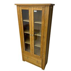 Light oak bookcase display cabinet, projecting square cornice over glazed double doors enclosing three adjustable shelves, single drawer base, on stile supports