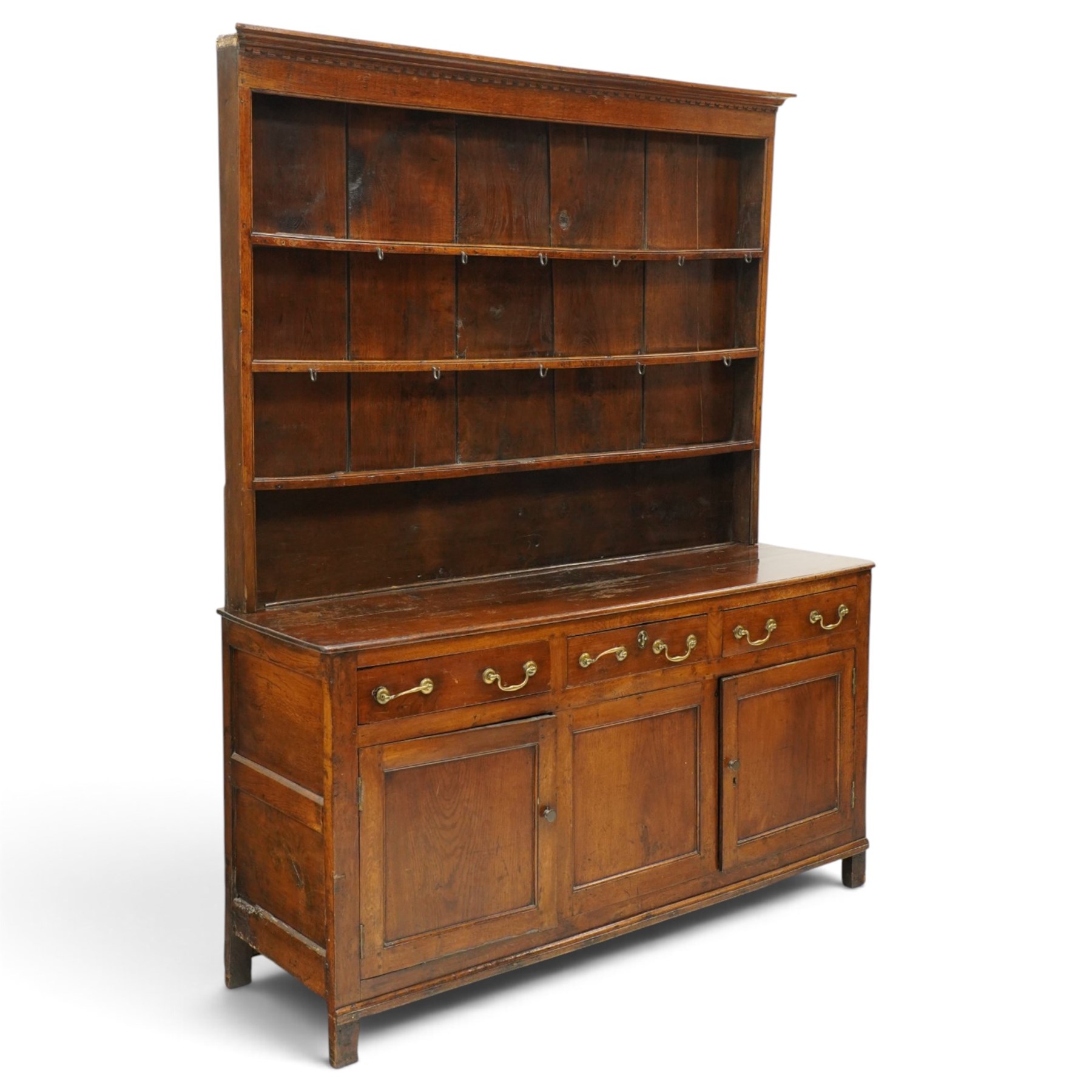 18th century oak dresser with rack, the three-tier plate rack with dentil-carved cornice, the plate shelves with moulded edge and wrought metal hooks, the base fitted with three drawers with brass swan-neck handles over three panelled cupboards, lower moulded edge on stile supports