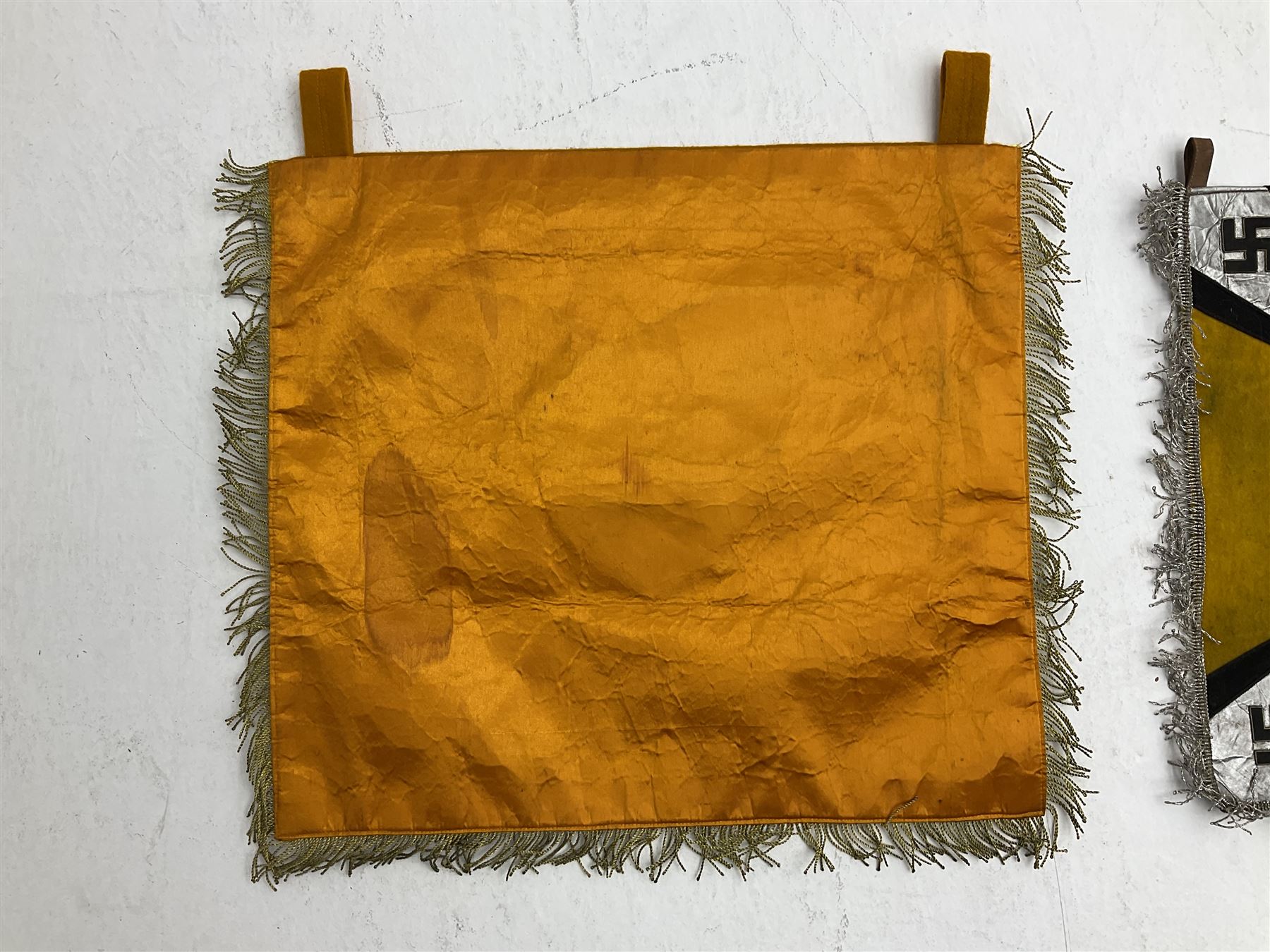 Two replica German Luftwaffe trumpet banners, one heavily silver wire embroidered with Luftwaffe eagle in a wreath, on an amber, white and black rayed background, , with silver wire tasselled border and four leather hanging loops, approx. 50 x 49cm; the other slightly larger heavily gold wire embroidered with the Luftwaffe eagle on a plain amber ground with gold wire tasselled border and two hanging loops (2)