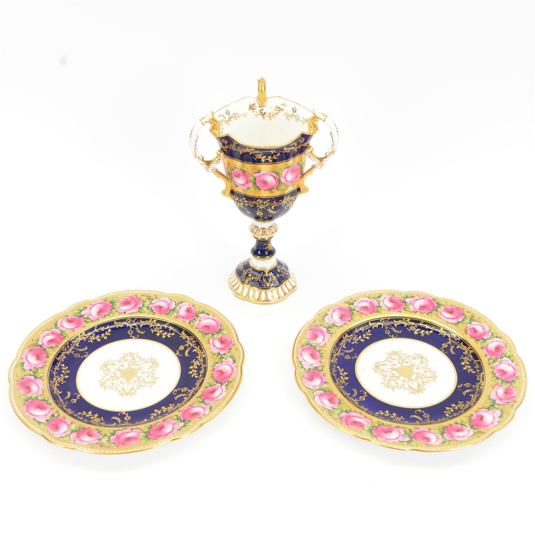 Early 19th century Coalport three handled goblet vase, shape no 114, and pair of plates, each hand painted with flowering pink roses upon a gilt band, against a dark blue ground heightened with trailing gilt tendrils, each marks beneath Coalport Swansea Period 1818, including handles vase H22.5cm, plates D23cm