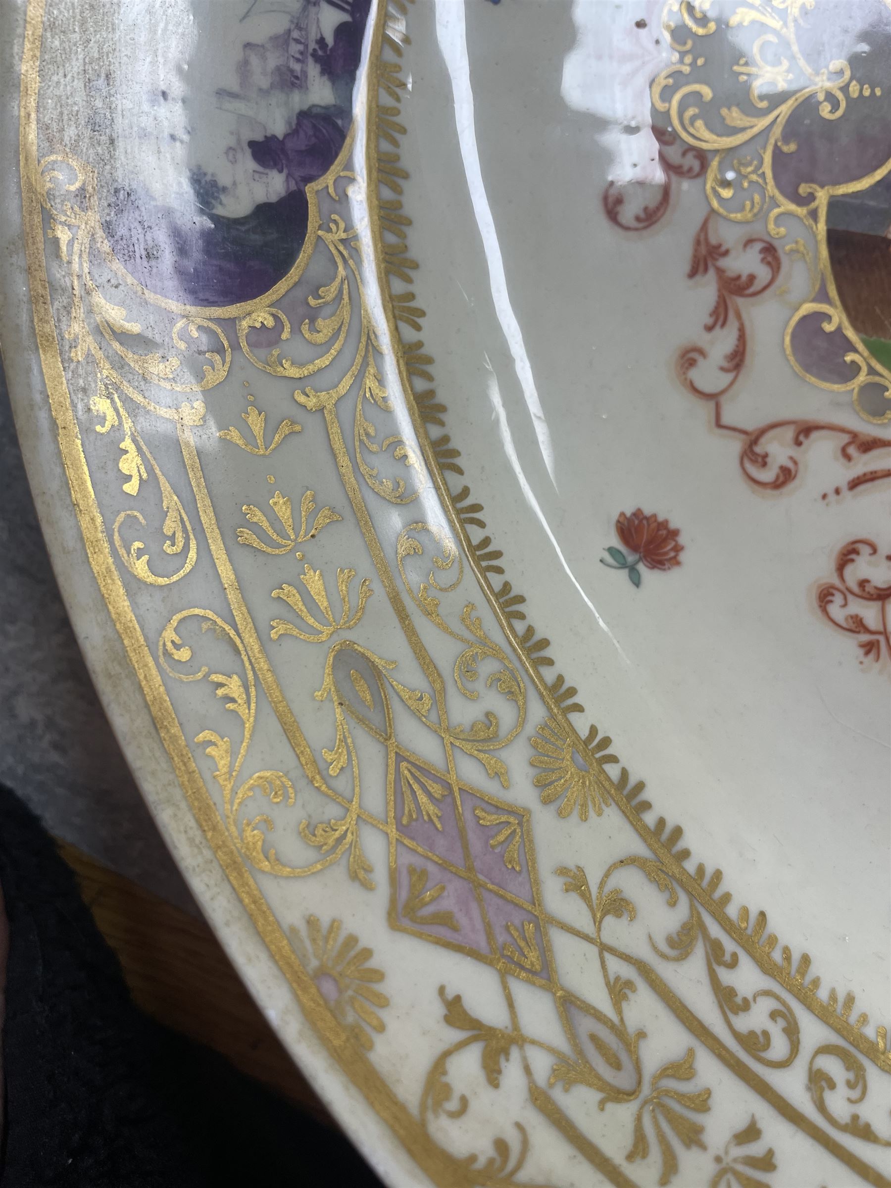 Large and impressive 18th century Meissen dish, finely painted with a chinoiserie scene to a central shaped reserve, depicting figures and attendants engaged in various pursuits, the reserve with gilt scrollwork edge, filled with Böttger lustre and detailed with iron red scrollwork, surrounded by flower heads, the rim with four quatrofoil Kauffahrtei scene panels of merchants and figures by a quayside in purple monochrome, with gilt scroll border, with under glazed blue crossed swords mark, and gilt gilders mark, D39cm
