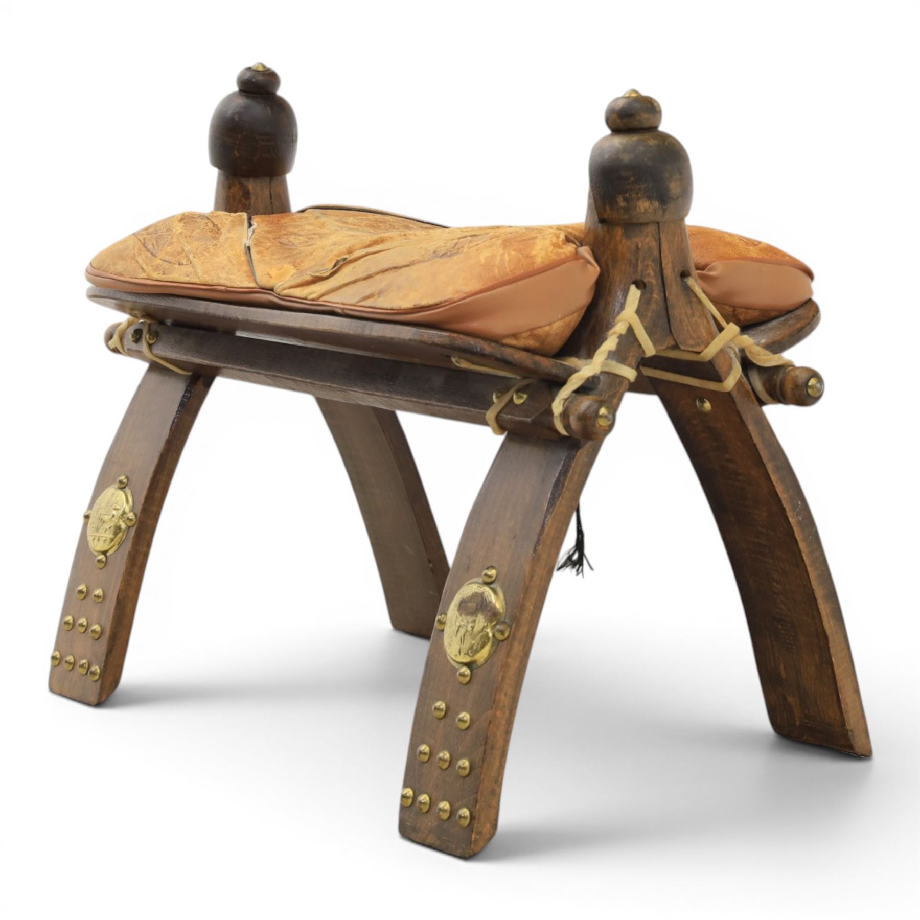 20th century Camel stool, with wooden turned finials, above a tooled leather seat, curved supports with brass applications 