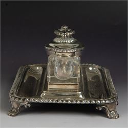 George IV silver desk stand, of rectangular form with rounded corners and oblique gadrooned rim, upon four embossed pad feet, with central pedestal dish of part fluted form with domed lid engraved with crest and with bud finial, with two recesses either side housing faceted glass inkwells, each with silver collars, hallmarked John & Thomas Settle, Sheffield 1825 & 1827, H11cm