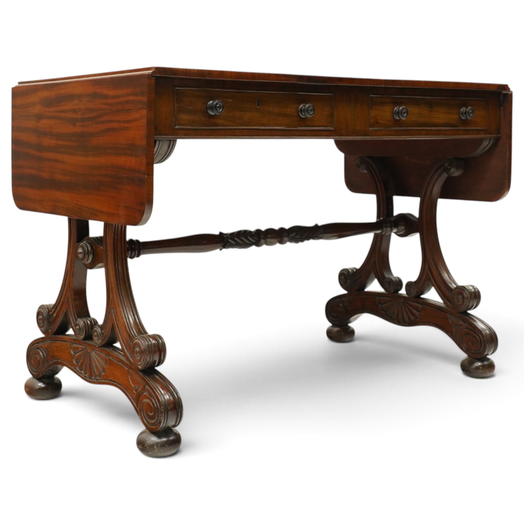 Fine 19th century figured mahogany sofa table, the rectangular twin drop-leaf top with rounded corners above two cock-beaded frieze drawers with opposing faux drawers, fitted with turned handles, raised on twin reeded and C-scroll-carved end supports with palmette-carved platform bases and compressed bun feet, united by a turned and foliate-carved stretcher