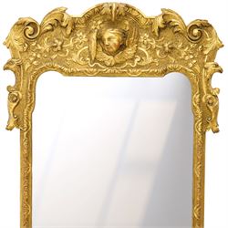 In the manner of Thomas Johnson - circa 1900 Georgian Revival giltwood wall mirror, the shaped rectangular plate within a foliate and scallop carved slip and richly ornamented frame, surmounted by a pierced cartouche crest centred with a protruding relief carved winged cherub mask and issuing scrolling acanthus and floral ornament, the sides with eagle masks over acanthus scrolls, terminating in a shaped apron with moulded rosette and spiralling acanthus leaves