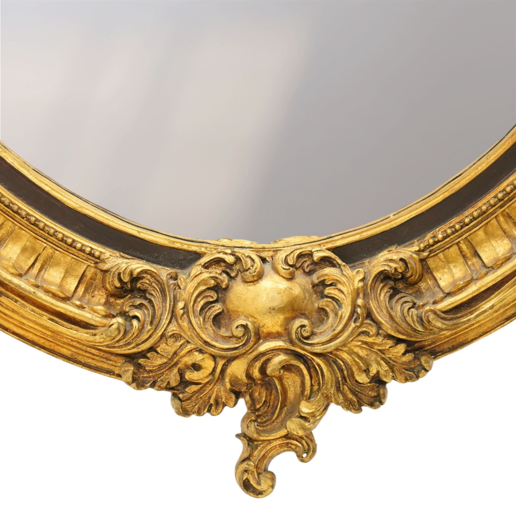 Pair of Rococo Revival gilt framed oval wall mirrors, the exuberant pediment formed of scrolling acanthus leaves and rocaille ornament, the oval bevelled plate set within with ebonised slip with outer repeating lappet decoration, over a rocaille cartouche terminal