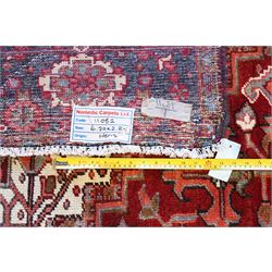 North West Persian Heriz hand-knotted carpet, brick red field with a large central angular star medallion with pendants surrounded by stylized geometric palmettes, serrated leaves and tribal motifs,, ivory corner spandrels, field enclosed by a dark indigo main border of stylize rosettes and angular vine motifs between multiple guard stripes, with cotton fringe ends 