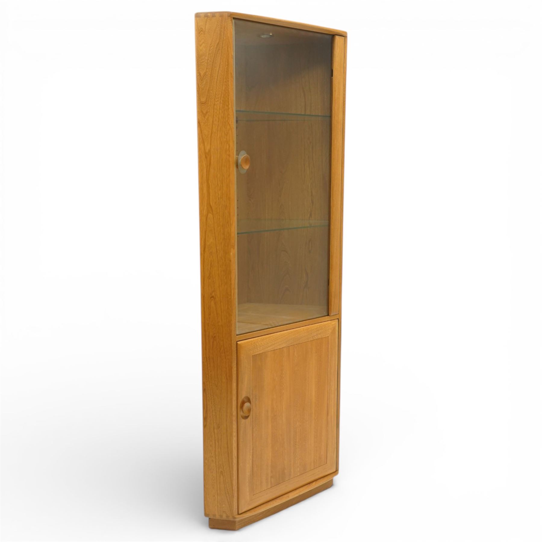 Ercol - elm corner display cabinet, Model 3856. Glazed upper section with glass door enclosing two glass shelves and interior light, above panel cupboard door, with rounded uprights and circular turned handles, on plinth base